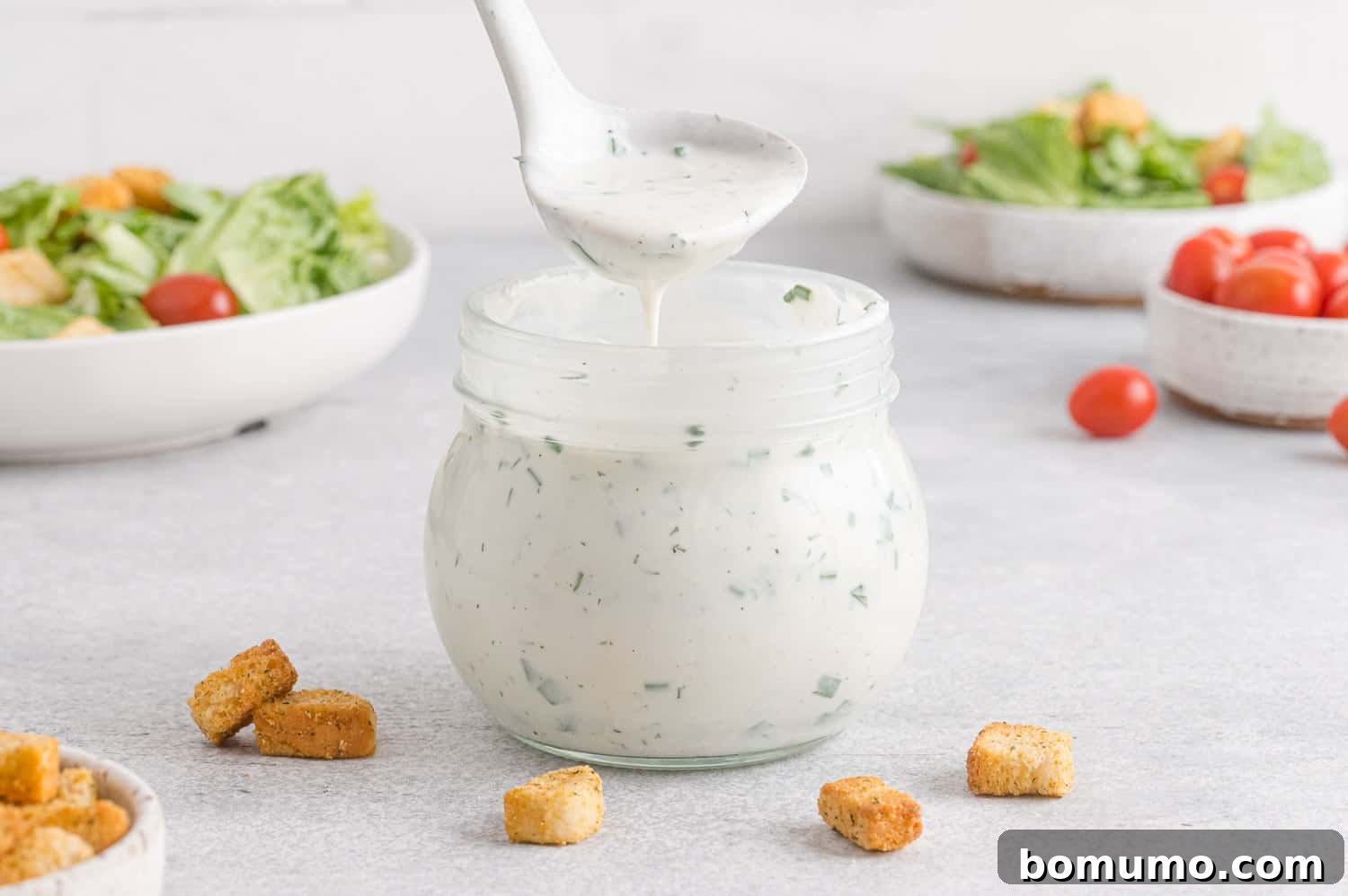 A close-up of healthy ranch dressing on a ladle, with a jar of dressing and two fresh salads blurred in the background, ready to be served.