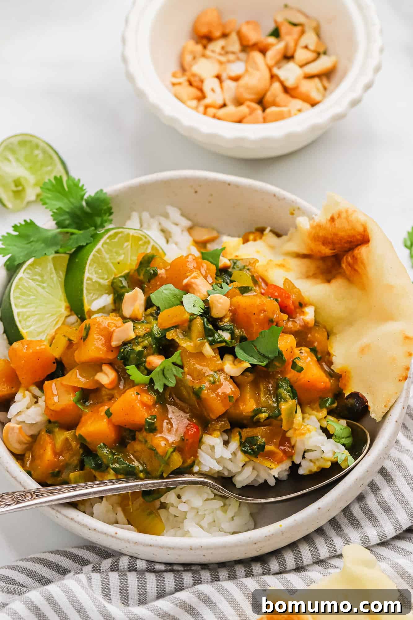 A beautifully presented bowl of butternut squash curry with a spoon, garnished with fresh cilantro and lime wedges, next to a portion of rice.