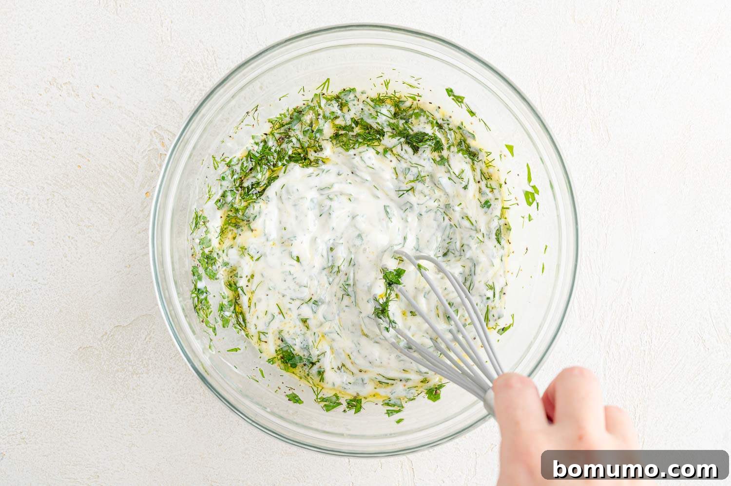 A whisk actively mixing the creamy dressing ingredients in a bowl until smooth.