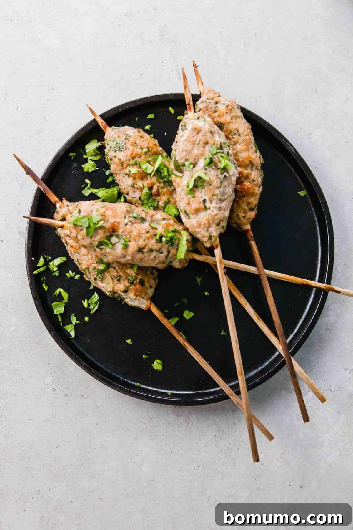 Overhead shot of several golden-brown chicken kofta skewers arranged artfully on a sleek black round plate, garnished with a generous sprinkle of fresh parsley.