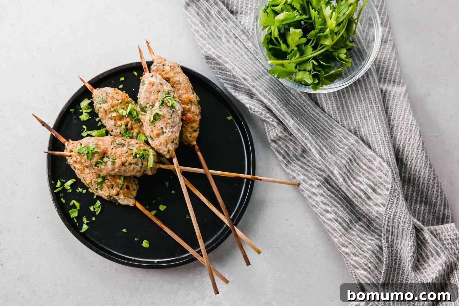 Overhead view of perfectly cooked chicken kofta on a serving plate, accompanied by a stylish gray striped cloth and a small bowl brimming with fresh parsley.