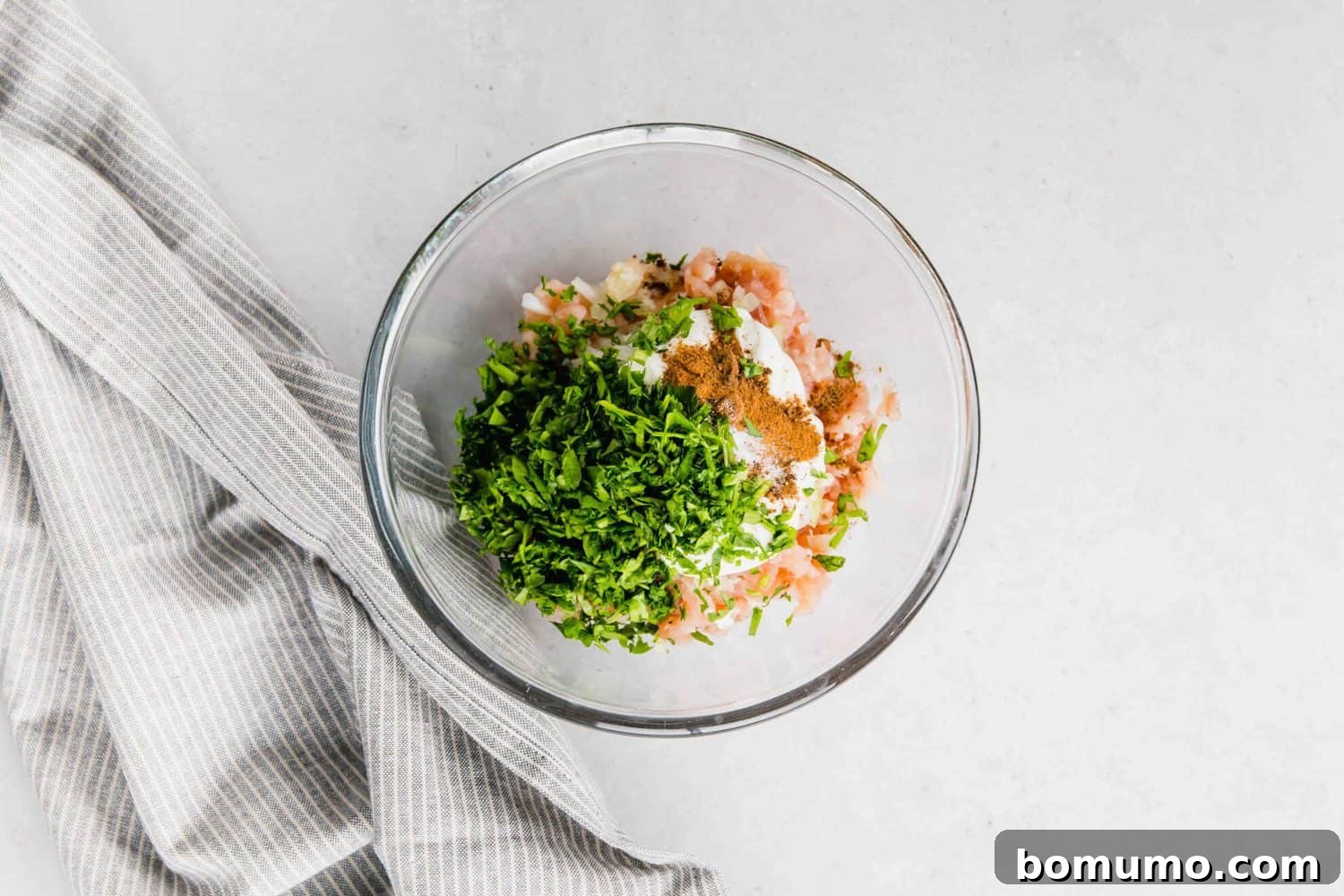 Overhead view of a clear glass mixing bowl containing all the fresh ingredients for Chicken Kofta before blending.