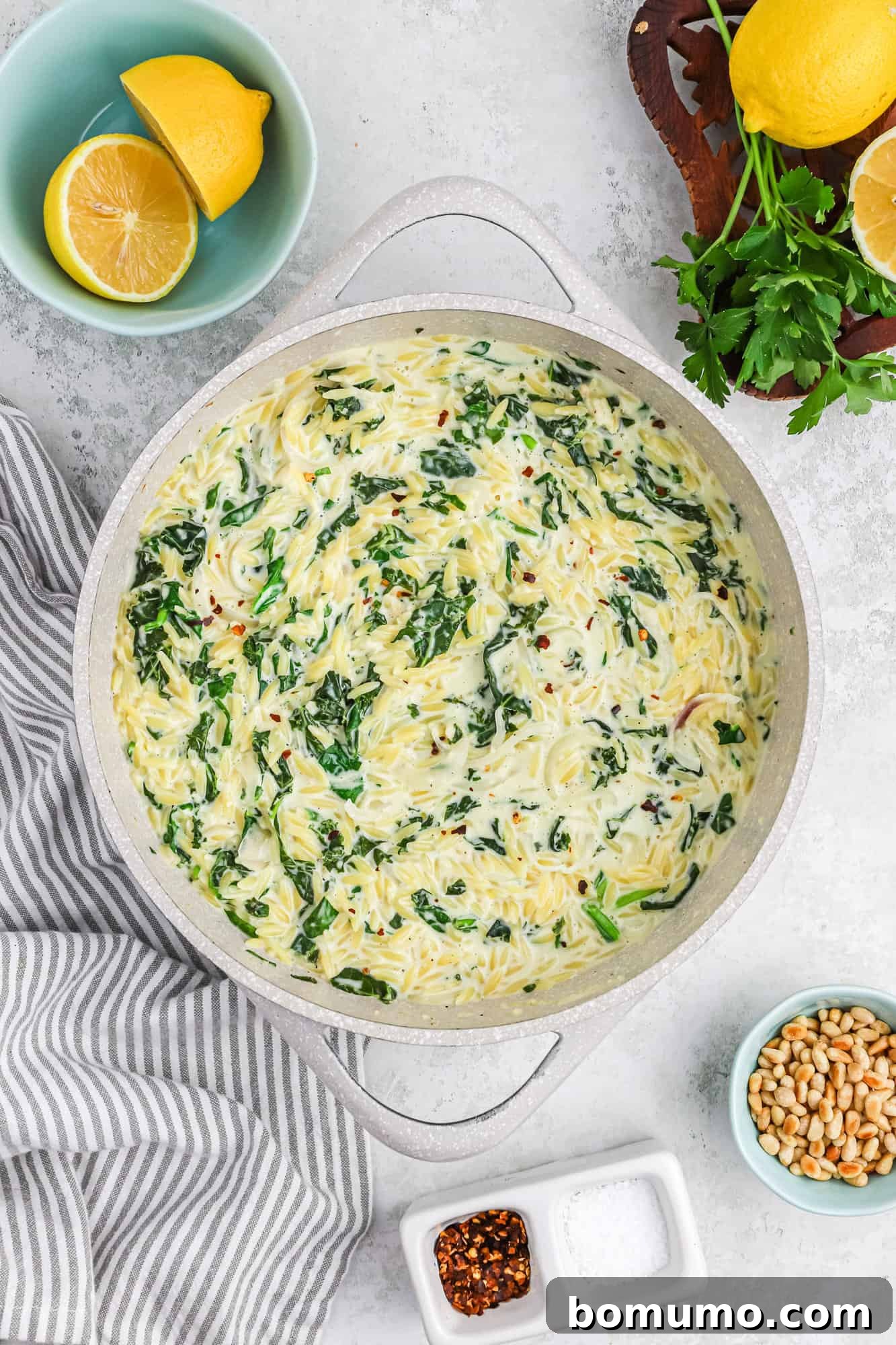 A large skillet filled with creamy orzo pasta, vibrant green spinach, and tender kale, ready to be served. Pine nuts add a delicate crunch on top.