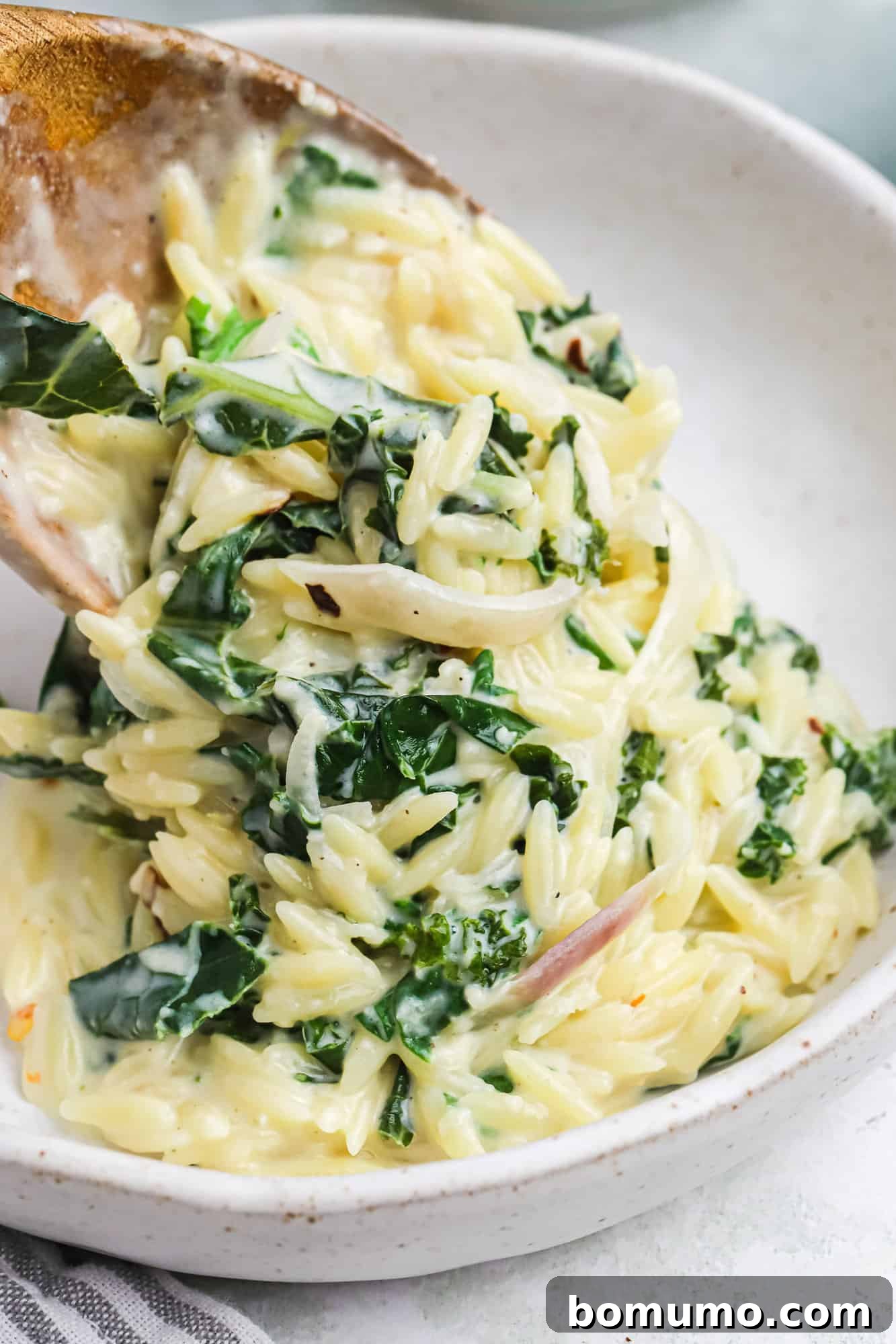 A serving spoon scoops a generous portion of creamy lemon orzo with spinach into a white bowl, showcasing the rich texture and vibrant greens.