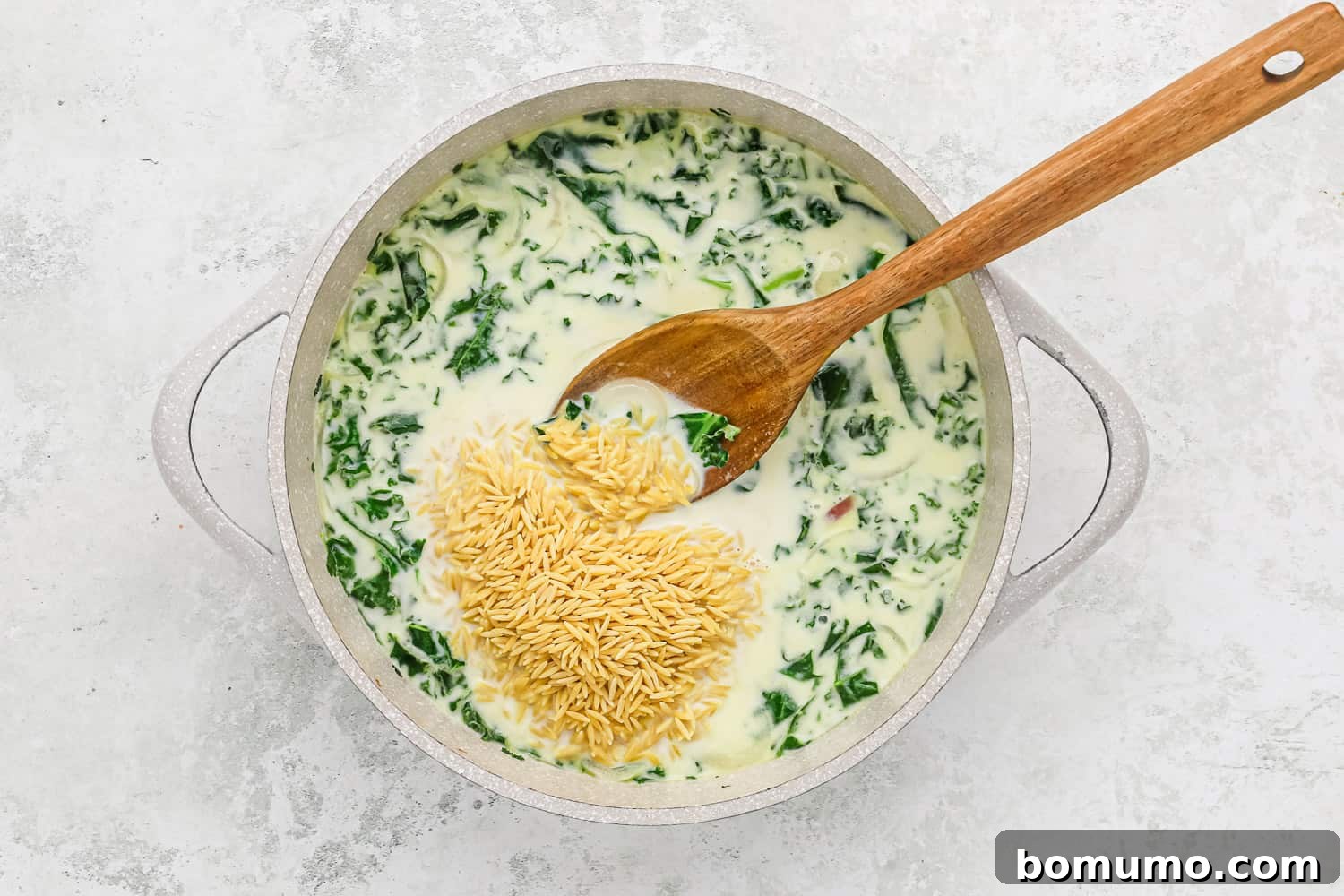 Uncooked orzo pasta is added to the simmering creamy broth and kale mixture in the skillet, ready to cook.