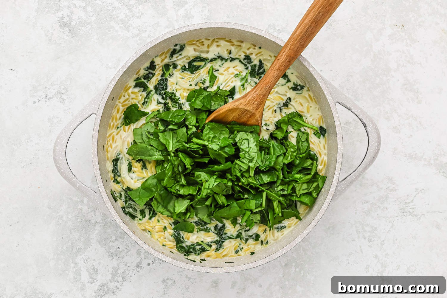 Fresh baby spinach leaves are stirred into the hot, creamy orzo and kale mixture in the pan, quickly wilting from the heat.