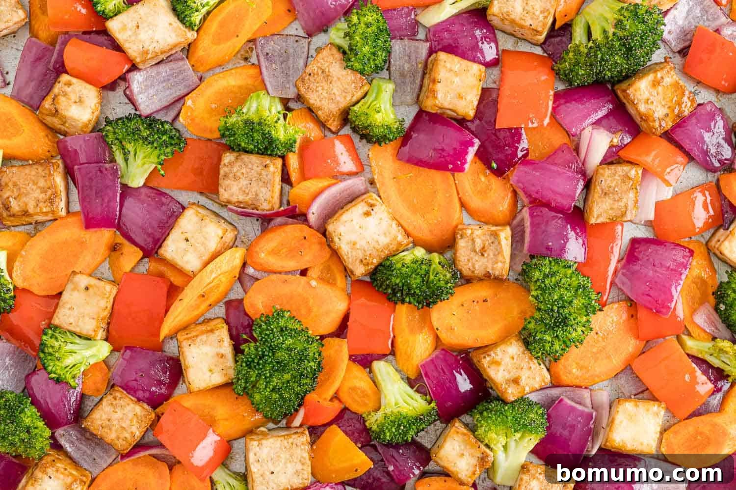A close-up view of oven-roasted tofu cubes, glistening and golden brown, mixed with tender-crisp vegetables on a sheet pan, ready to be sauced.