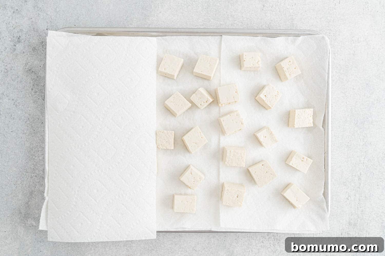 Paper towels carefully placed over cubed tofu on a sheet pan, initiating the pressing process.