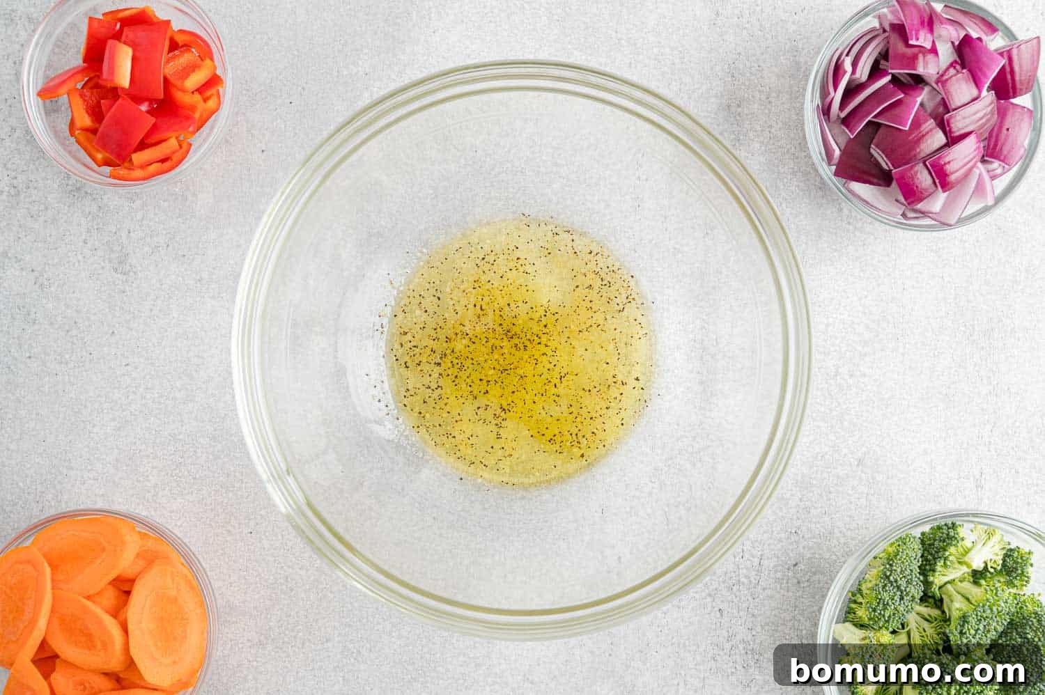 A large mixing bowl containing olive oil and various seasonings, surrounded by a colorful assortment of fresh vegetables, ready for tossing.