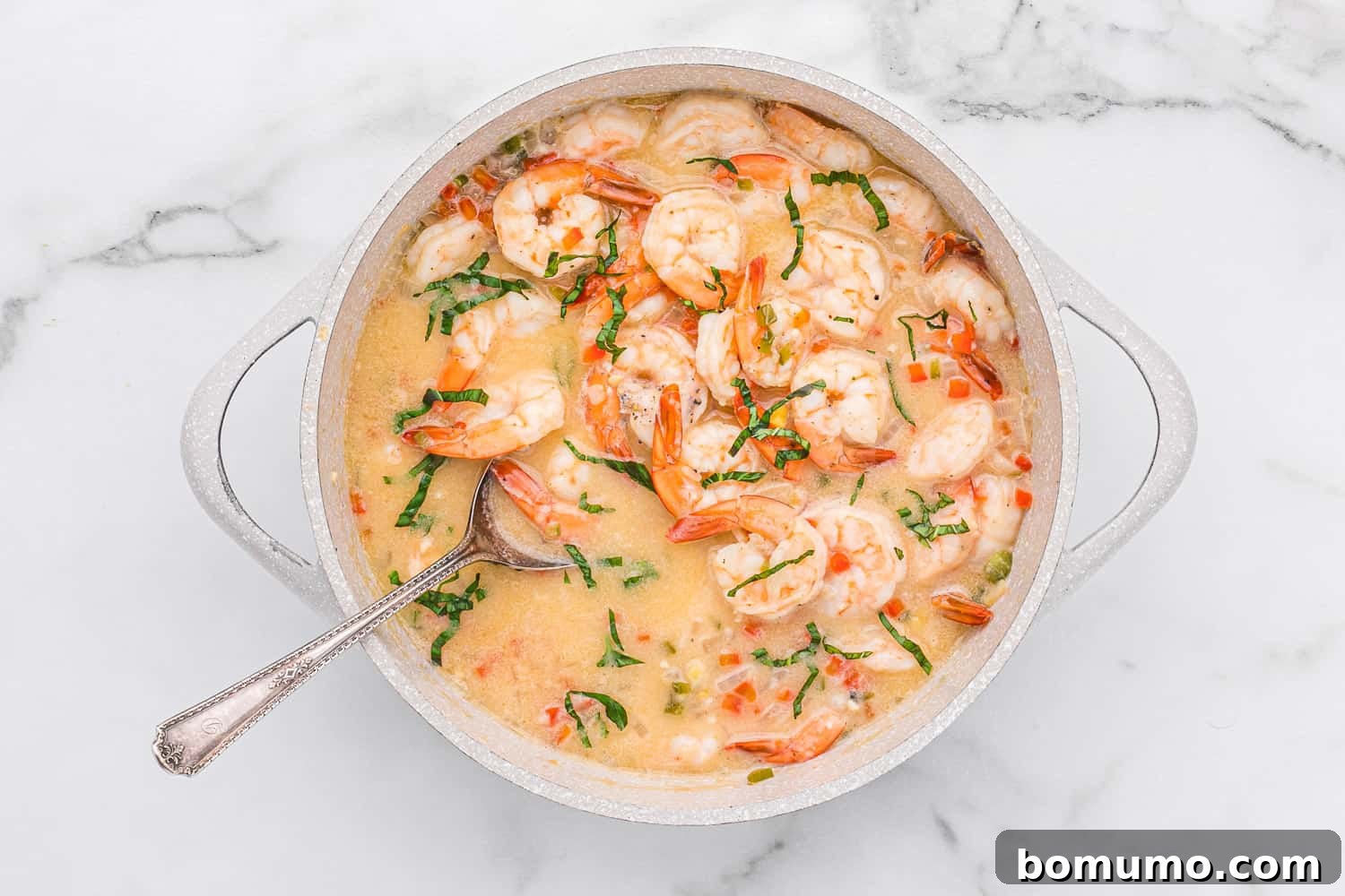 A pot of creamy coconut shrimp, garnished with fresh herbs and diced vegetables, served with a metal spoon.