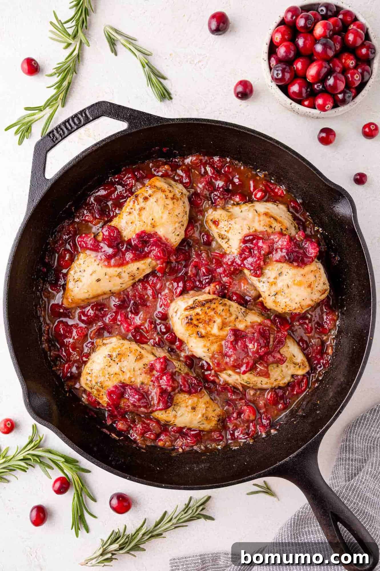 Cranberry Glazed Chicken 13 Skillet with cooked chicken breasts topped with cranberry sauce, surrounded by fresh cranberries and rosemary sprigs on a light surface.