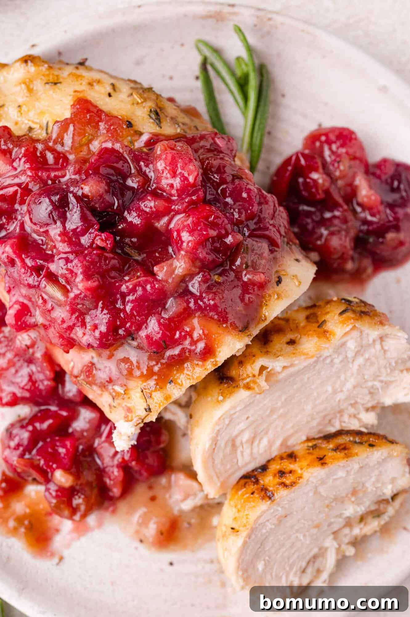 Cranberry Glazed Chicken 3 Cooked chicken breast topped with a chunky cranberry sauce, garnished with fresh rosemary on a white plate.