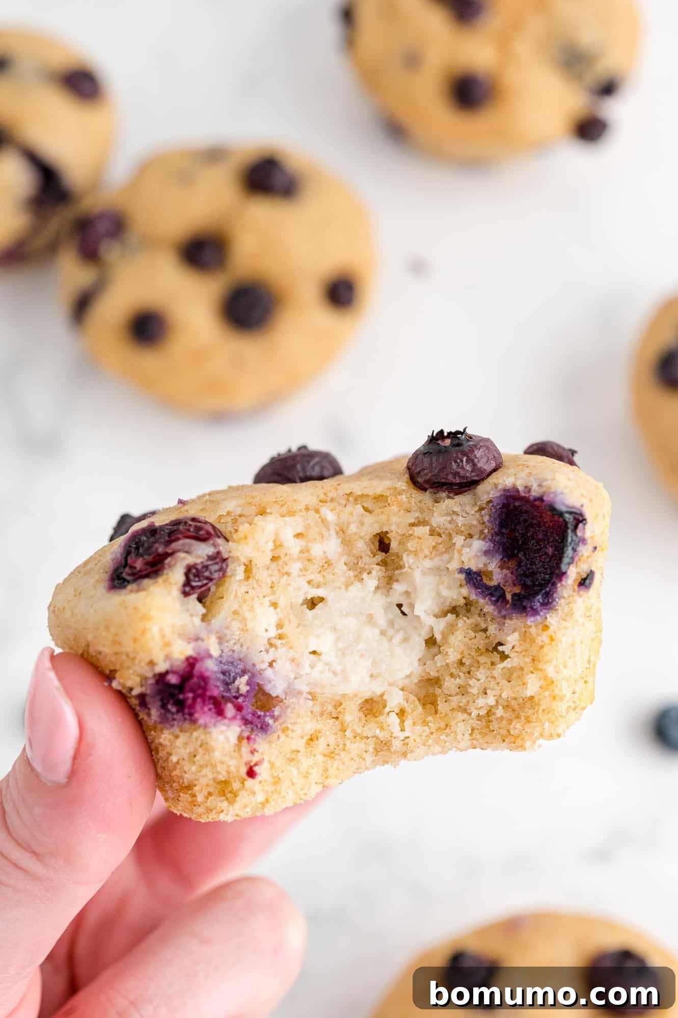 A freshly baked blueberry muffin with cream cheese filling, held delicately in a hand, showing its golden-brown exterior and inviting texture.