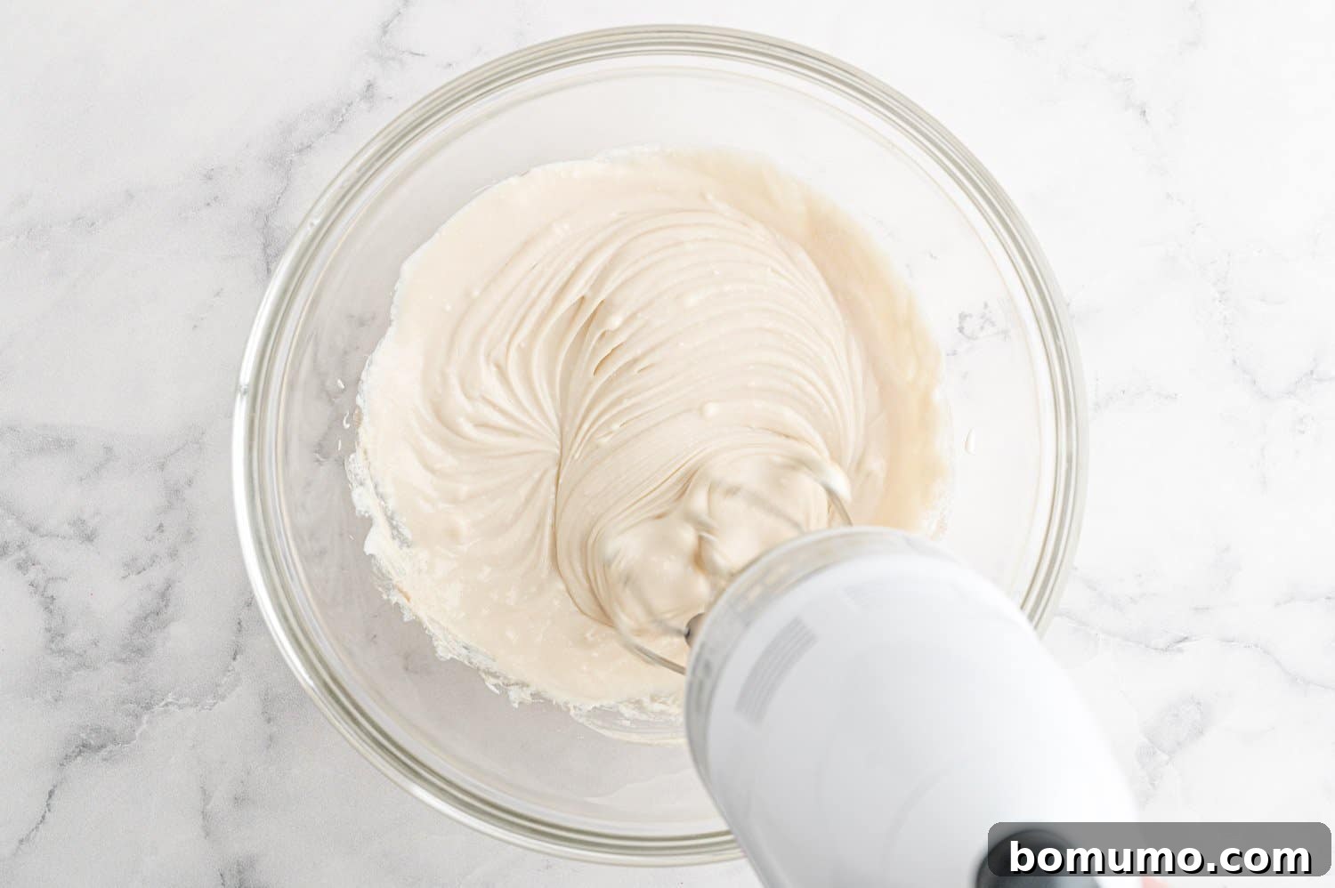 A bowl of smooth cream cheese mixture, with an electric mixer nearby, showing the filling ready for the muffins.