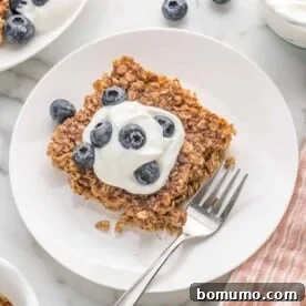 Square of baked oatmeal on a white plate, topped with yogurt and fresh blueberries.