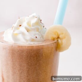A decadent chocolate milkshake adorned with a dollop of whipped cream and a banana slice on the rim, featuring a fun blue straw.