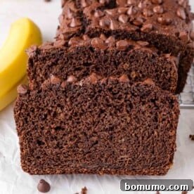 A close-up view of sliced healthy chocolate banana bread with melted chocolate chips, resting beside a fresh banana on a white surface.