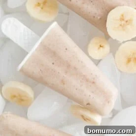 A refreshing banana popsicle stands amidst a scattering of fresh banana slices, highlighting its creamy texture.