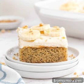 A slice of banana cake generously topped with white cream cheese frosting, fresh banana slices, and chopped nuts.