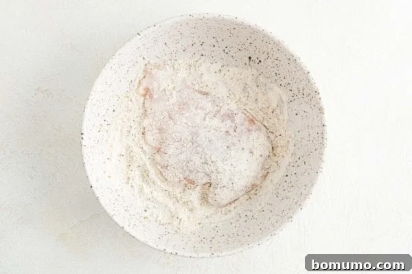 A chicken cutlet being dredged in a bowl of seasoned flour, ensuring an even coating.