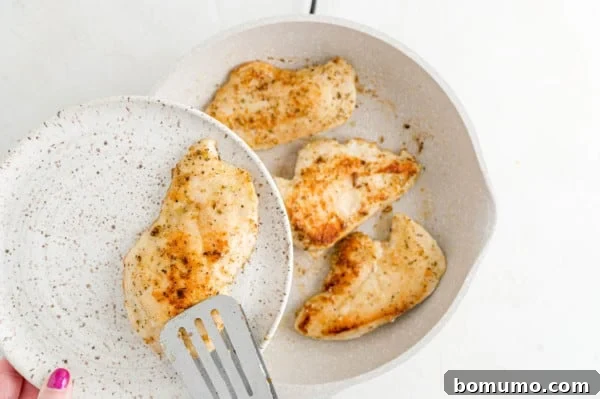 A plate holding a perfectly pan-fried chicken cutlet, with more chicken searing in the skillet below.