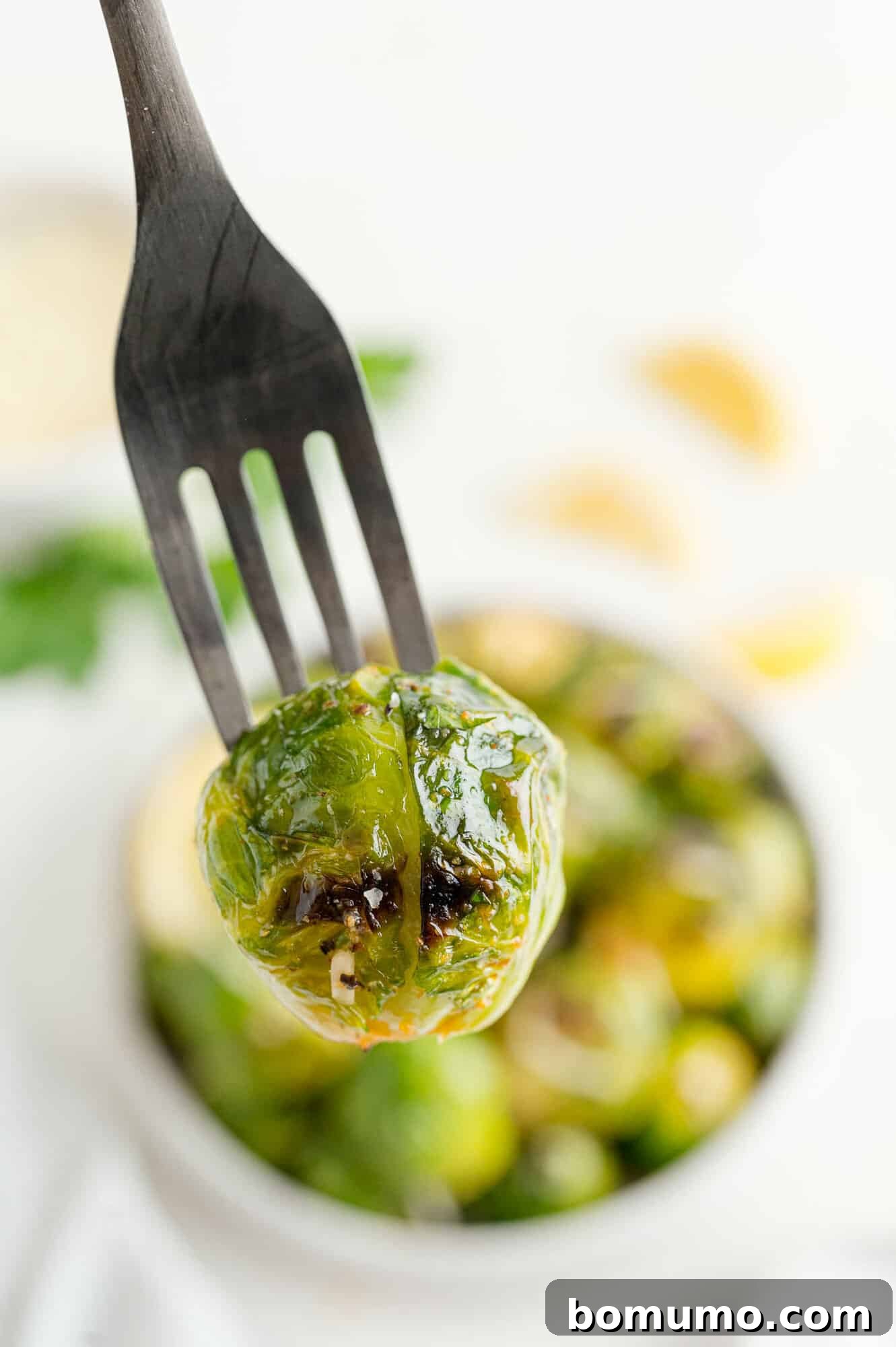 These are the best grilled Brussels sprouts! Cooked until golden and tossed in a combination of zesty lemon, parmesan, and fresh herbs. Close up of a grilled Brussels sprout on a fork, held over a bowl of Brussels sprouts in the background.