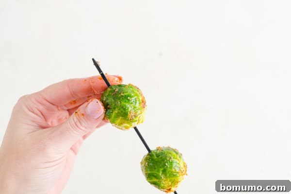 These are the best grilled Brussels sprouts! Cooked until golden and tossed in a combination of zesty lemon, parmesan, and fresh herbs. A hand adds seasoned Brussels sprouts onto a skewer.