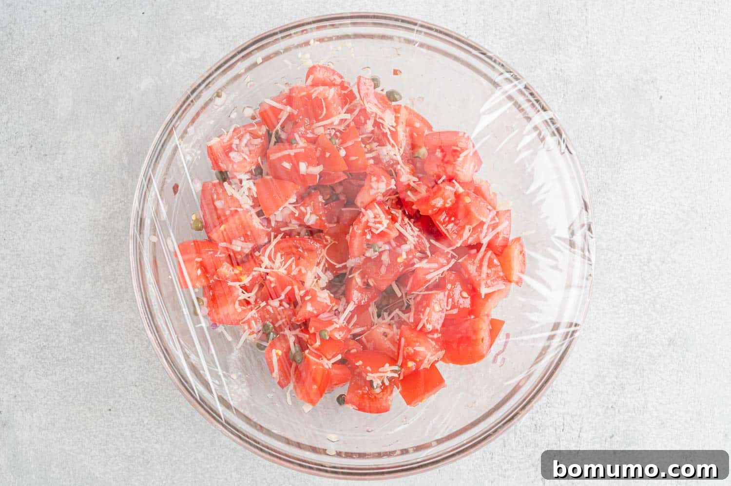 Tomatoes marinating in a glass bowl covered with plastic wrap.