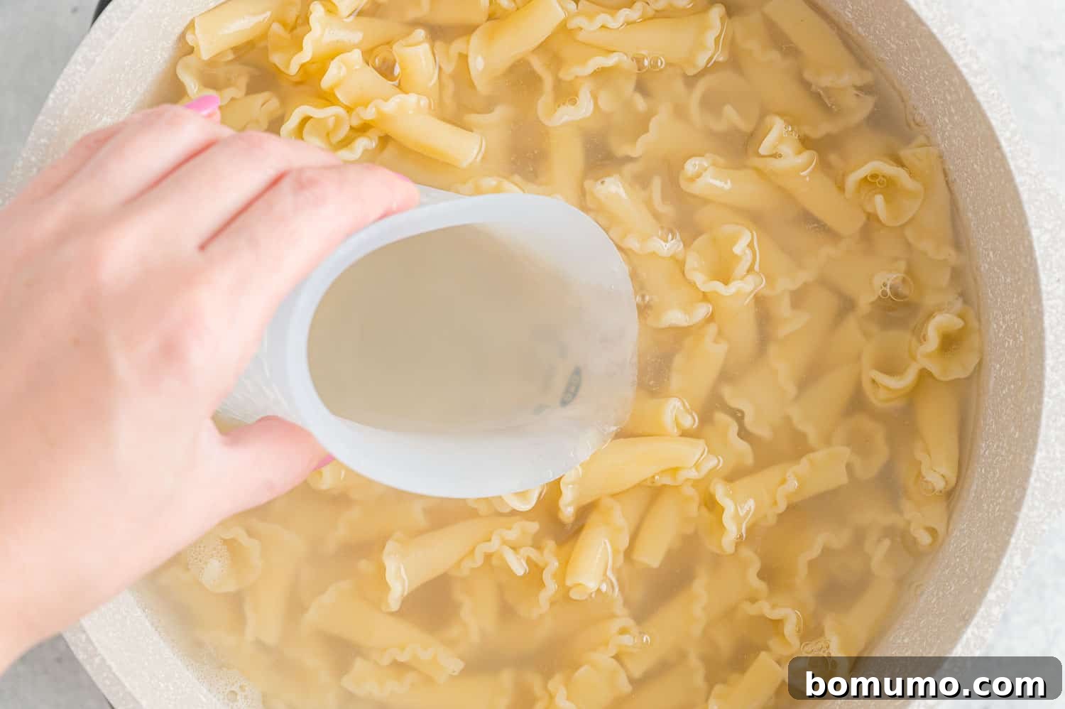 A hand scooping a cup of reserved pasta water from a pot of cooked pasta.
