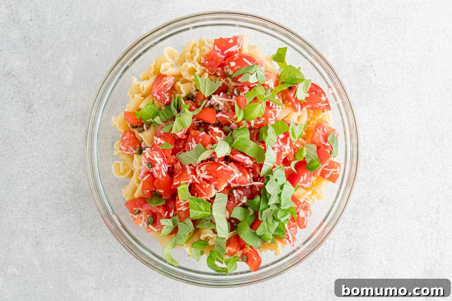 Fresh marinated tomatoes, basil, and seasonings added to a glass bowl with cooked pasta.