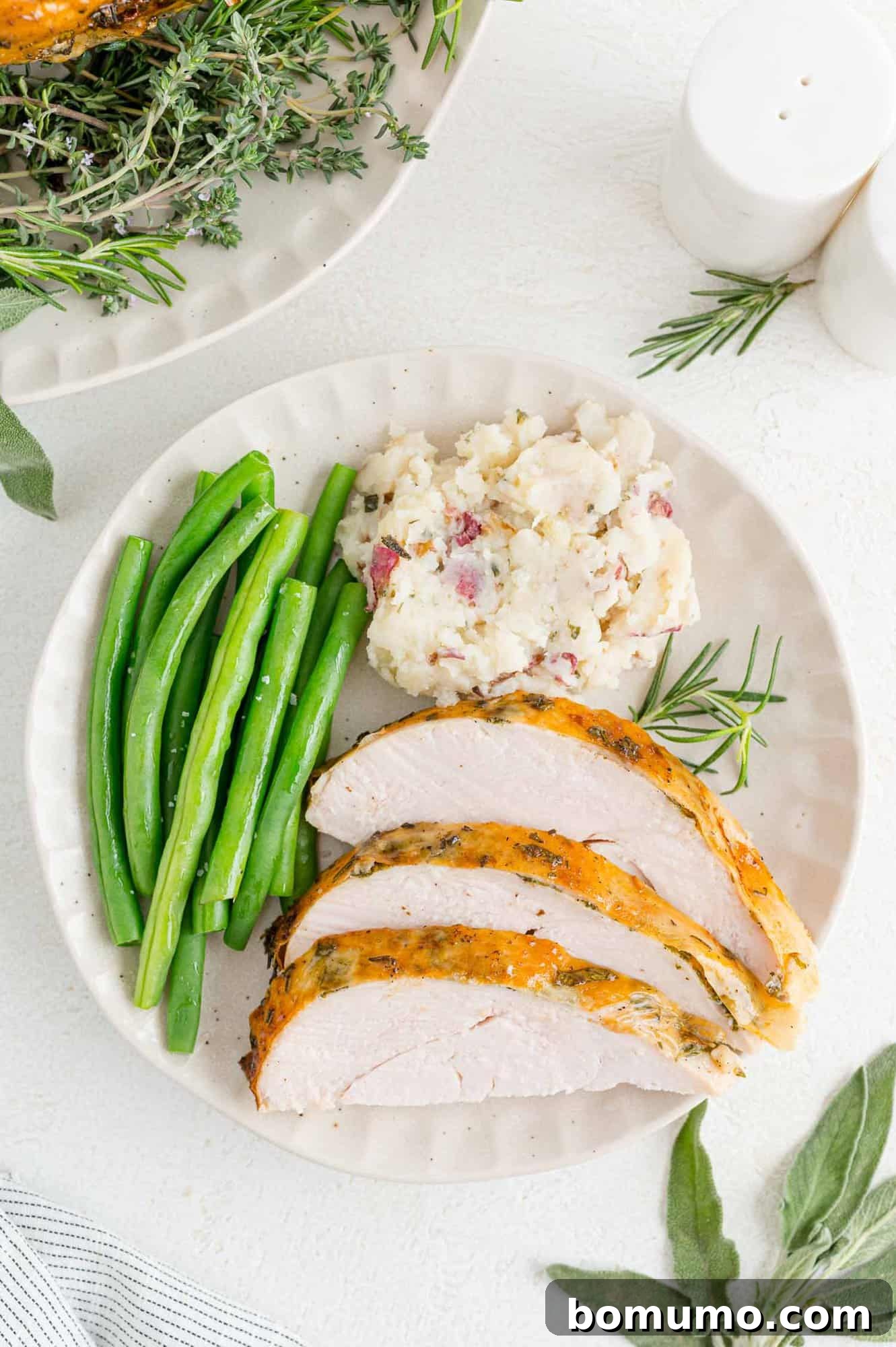 Three generous slices of juicy, herb roasted turkey breast served on a white plate, accompanied by a spoonful of creamy mashed potatoes and vibrant steamed green beans.