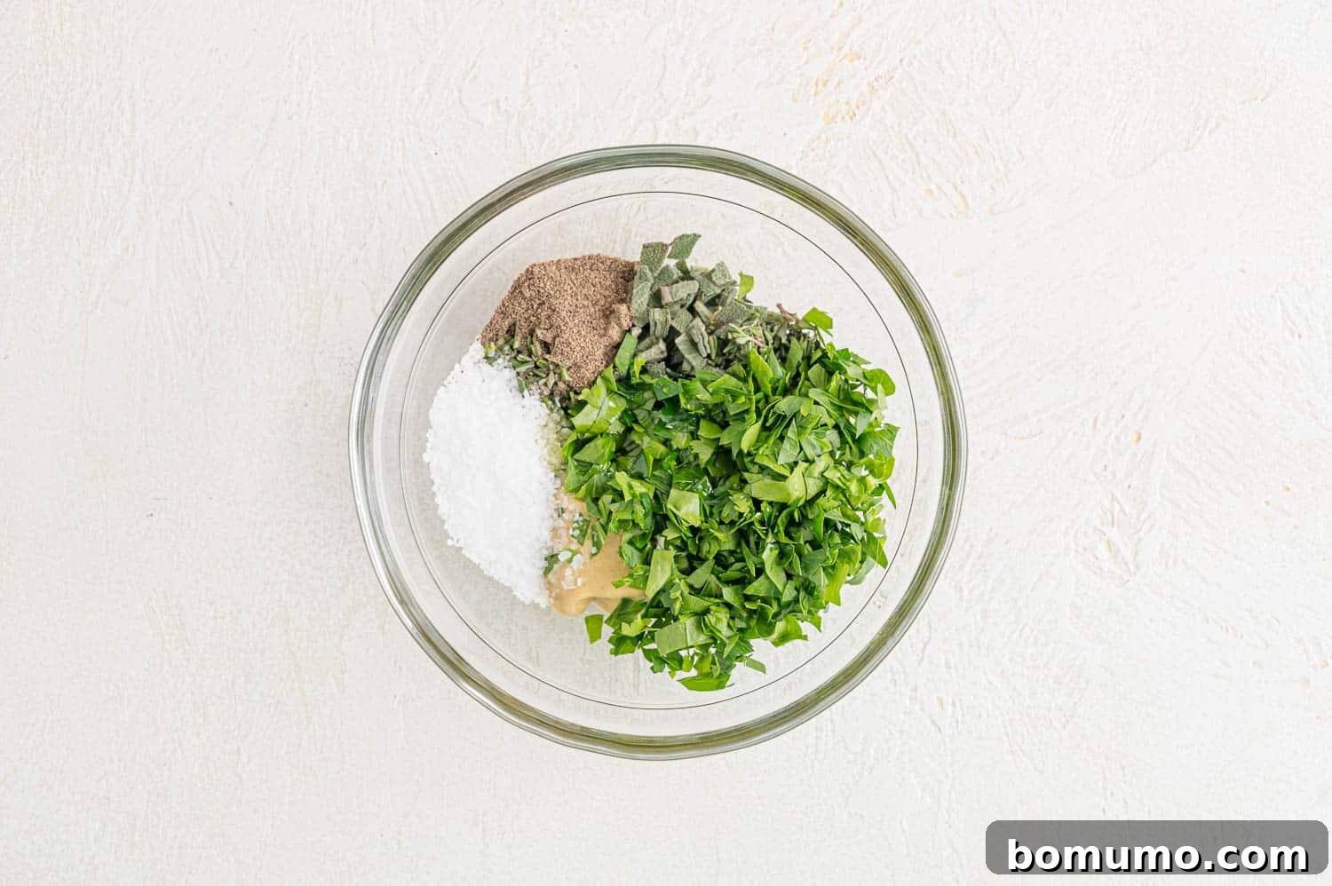 Freshly chopped herbs, minced garlic, Dijon mustard, salt, pepper, and olive oil waiting to be mixed in a small white bowl.