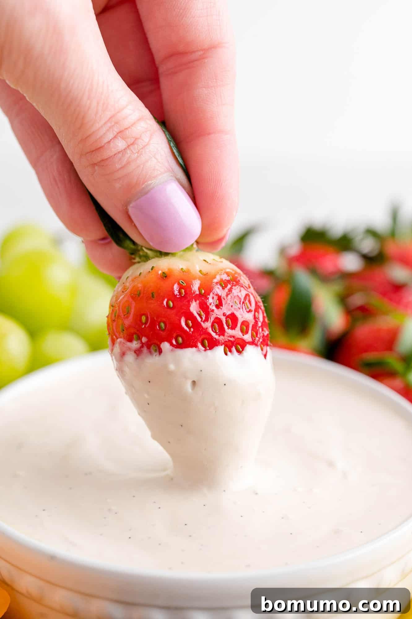 A hand dips a strawberry into a bowl of cream cheese fruit dip in the center of a fruit platter.