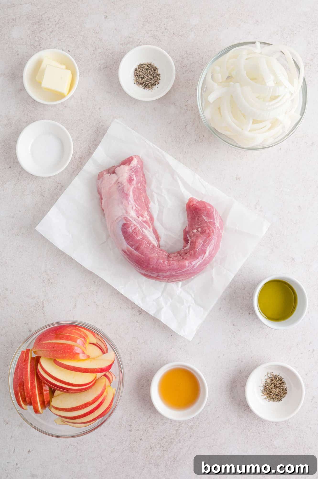 A collection of fresh ingredients laid out on a kitchen counter: pork tenderloin, vibrant apples, a large onion, butter, olive oil, and herbs.