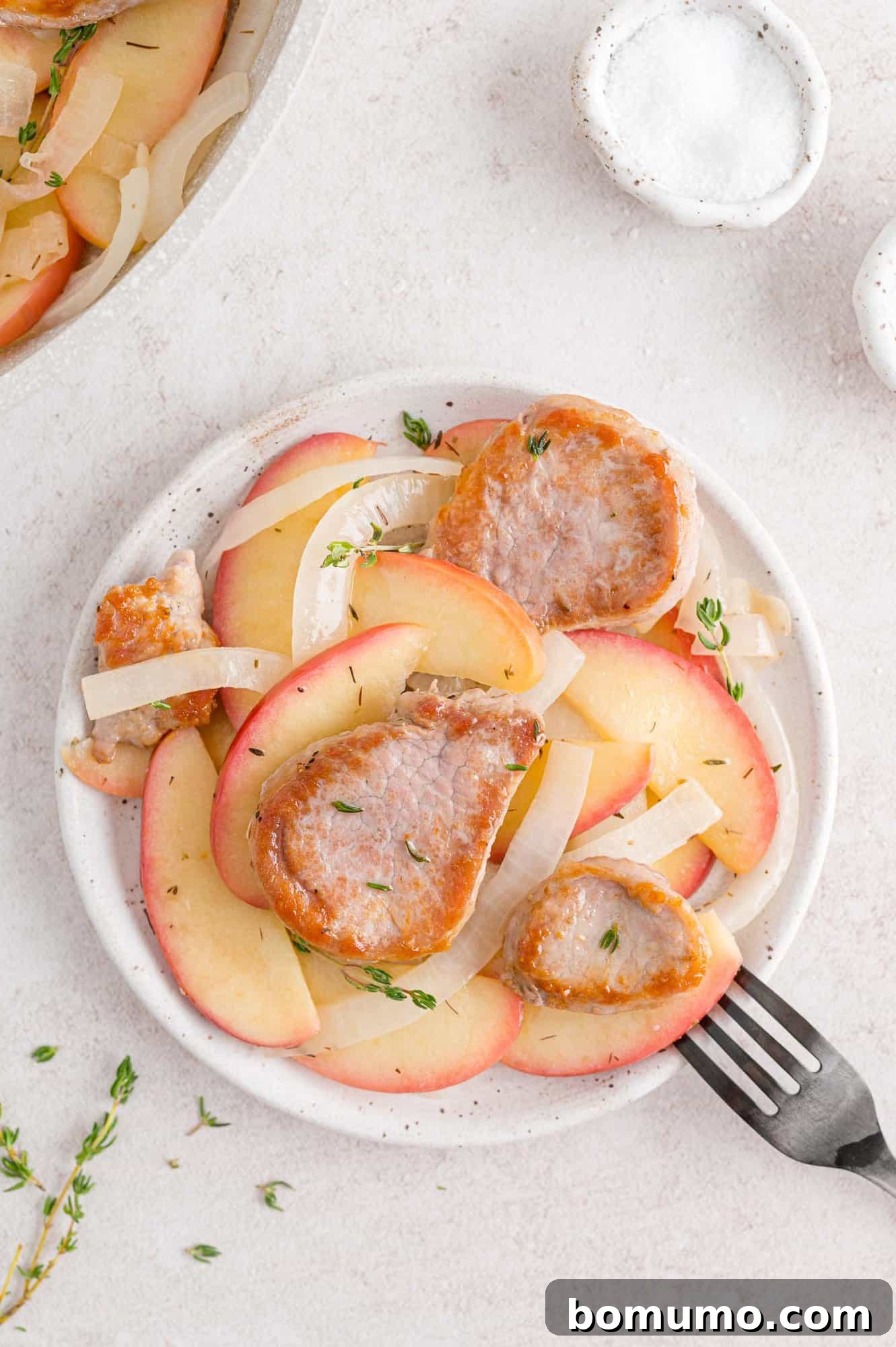 A perfectly plated dish of pork tenderloin slices, caramelized apples, and onions, garnished with fresh thyme, on a white plate.