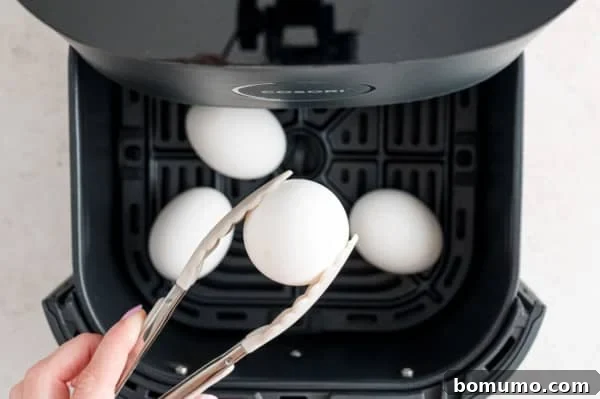 A pair of kitchen tongs carefully lifting a perfectly cooked boiled egg from the hot air fryer basket, demonstrating the ease and safety of removal.