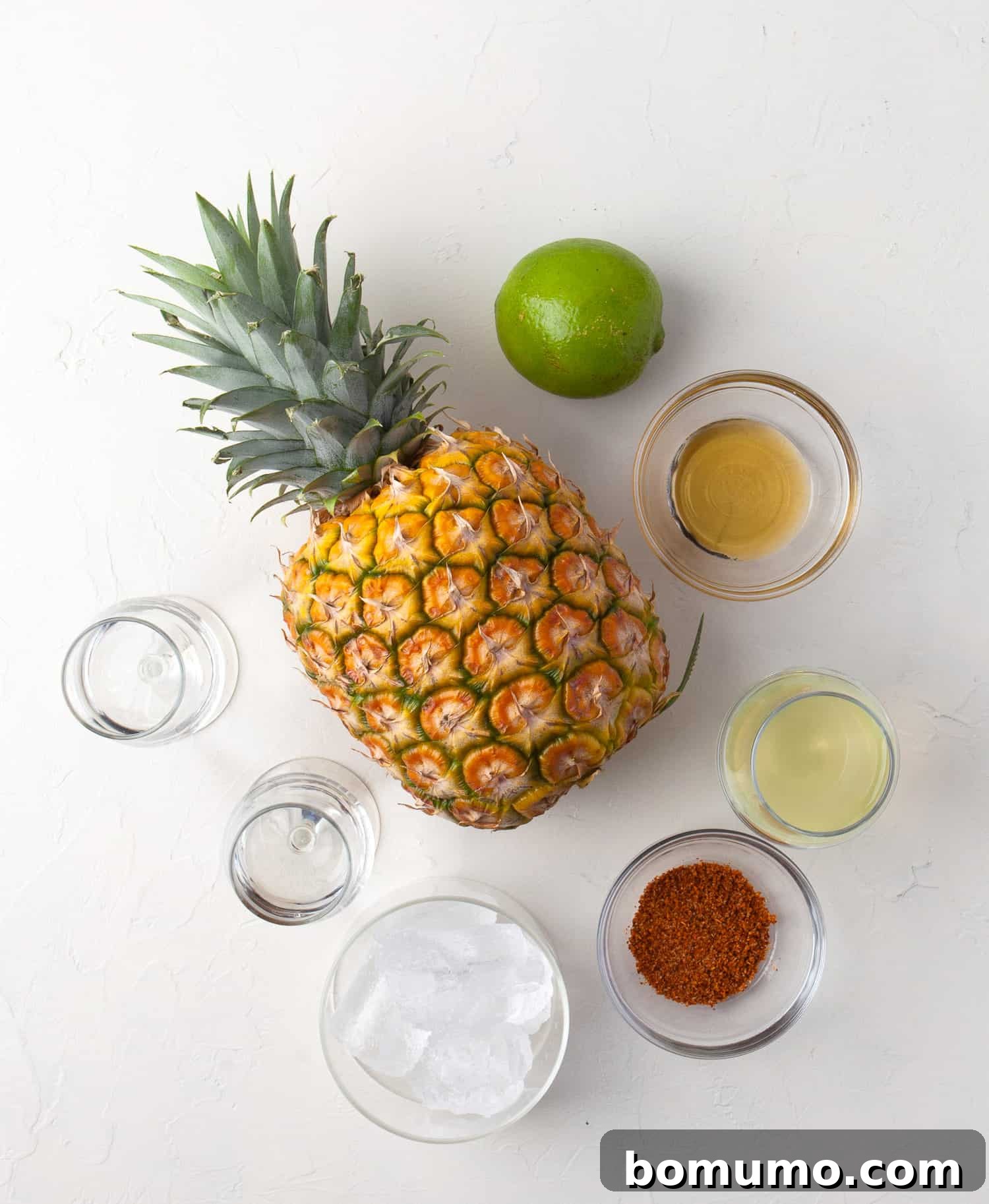 A collection of fresh ingredients laid out, including limes, a whole pineapple, a bottle of tequila, a bottle of triple sec, and agave syrup, ready for crafting a pineapple margarita.