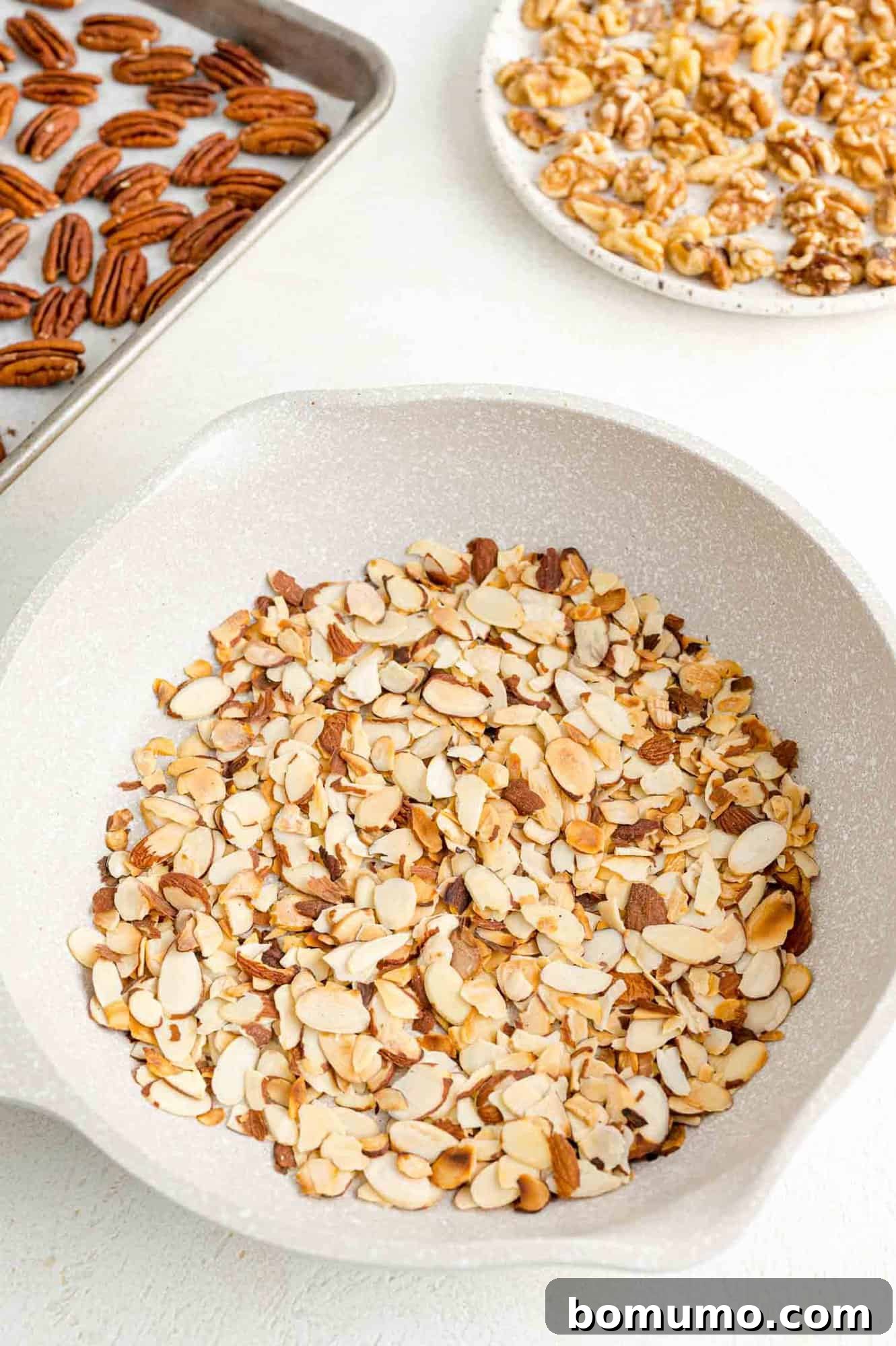 Toasting almonds is a great way to bring out their crunch and flavor. Learn how to toast almonds in the oven, microwave, and on the stovetop! Toasted almonds in a white skillet next to a tray of toasted pecans and a plate of toasted walnuts.