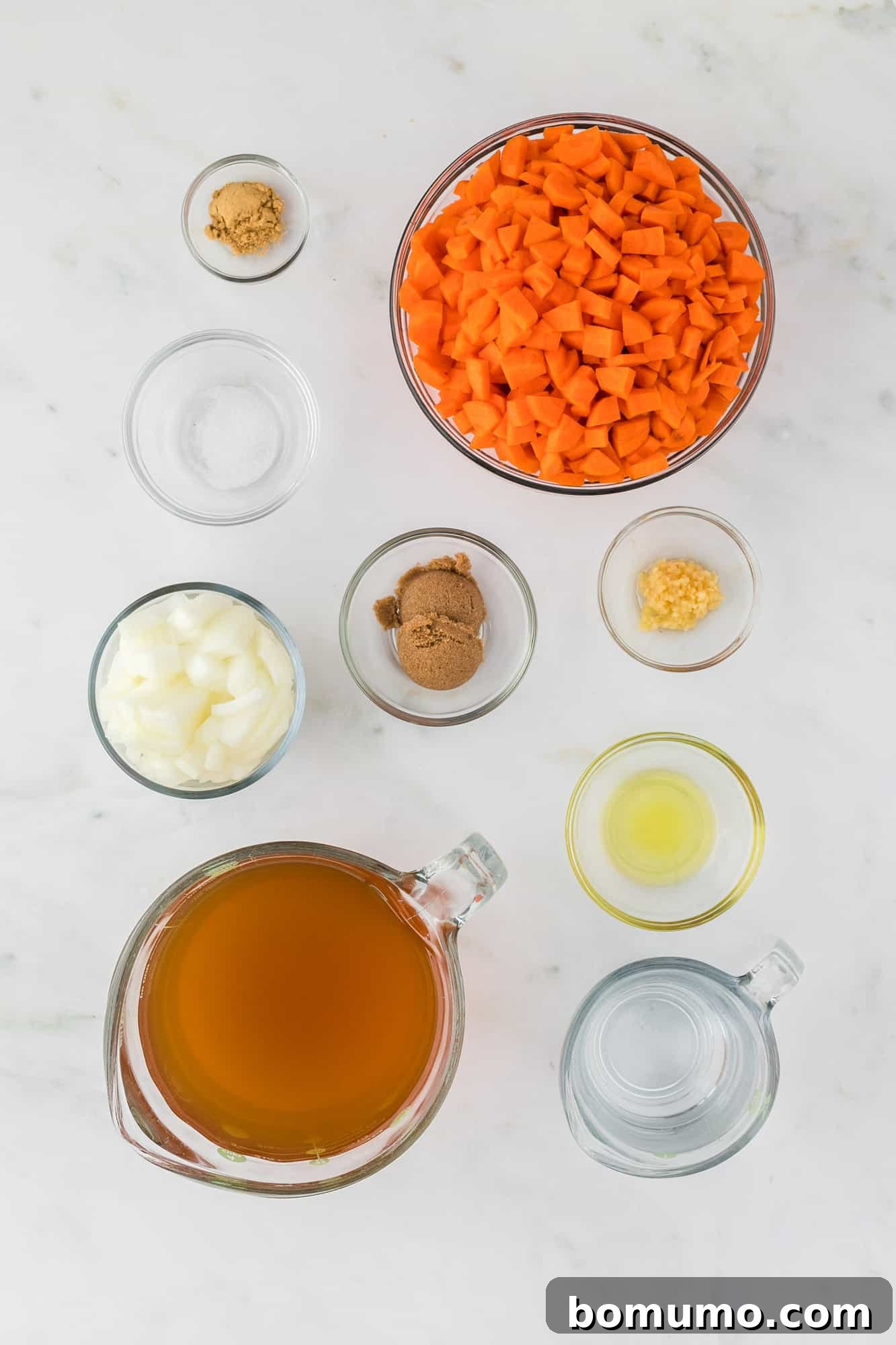 Overhead view of all individual ingredients for creamy carrot soup in separate white bowls, including carrots, onion, garlic, broth, cream, and spices.