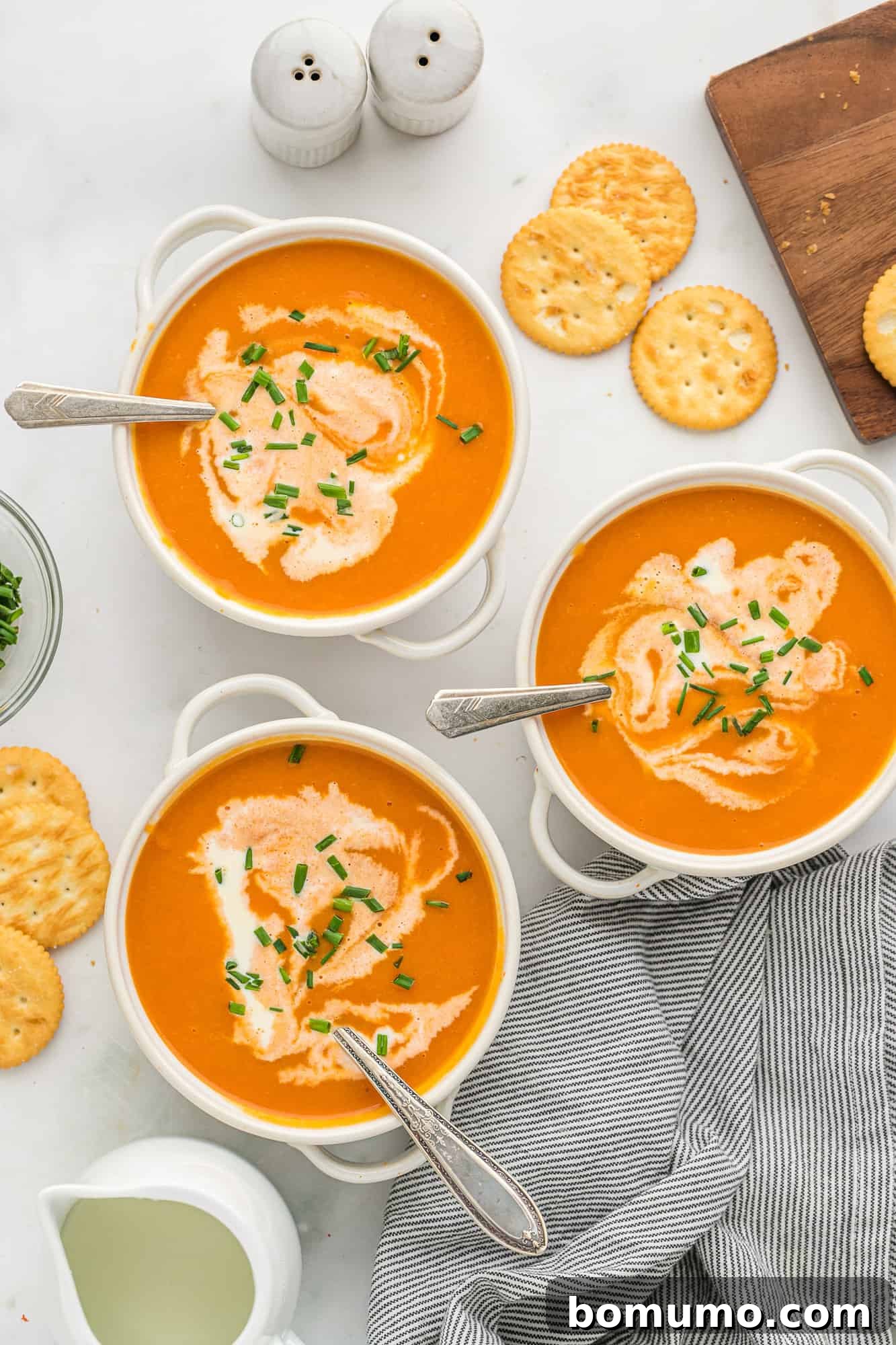 Three elegant bowls of creamy carrot soup, garnished with cream swirls and chives, with spoons resting in each.