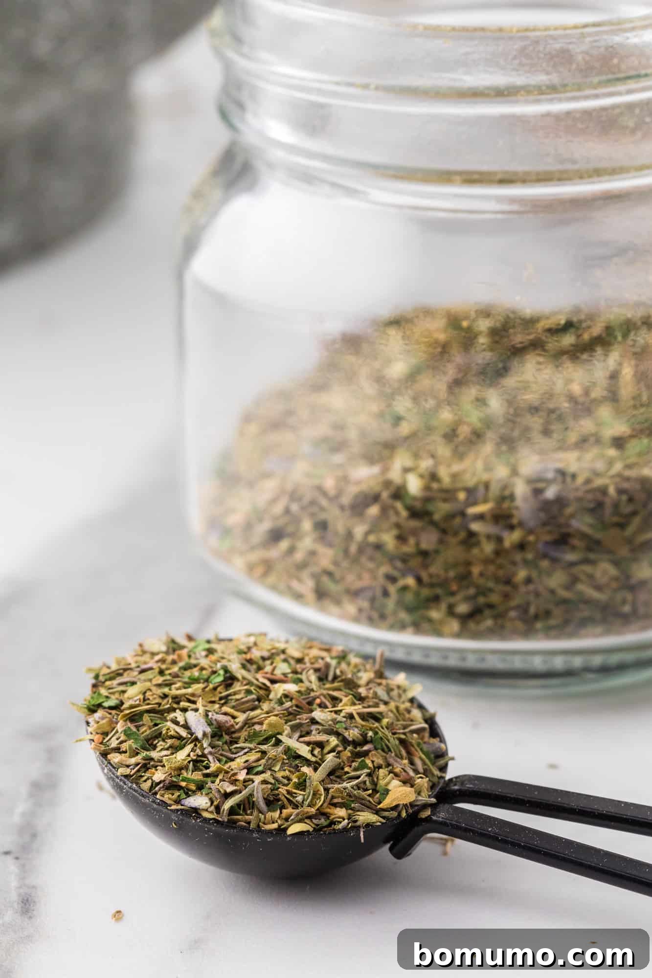 A small ceramic spoon holds a delicate mixture of Herbes de Provence, with a glass jar filled with the blend in the background, ready for use.