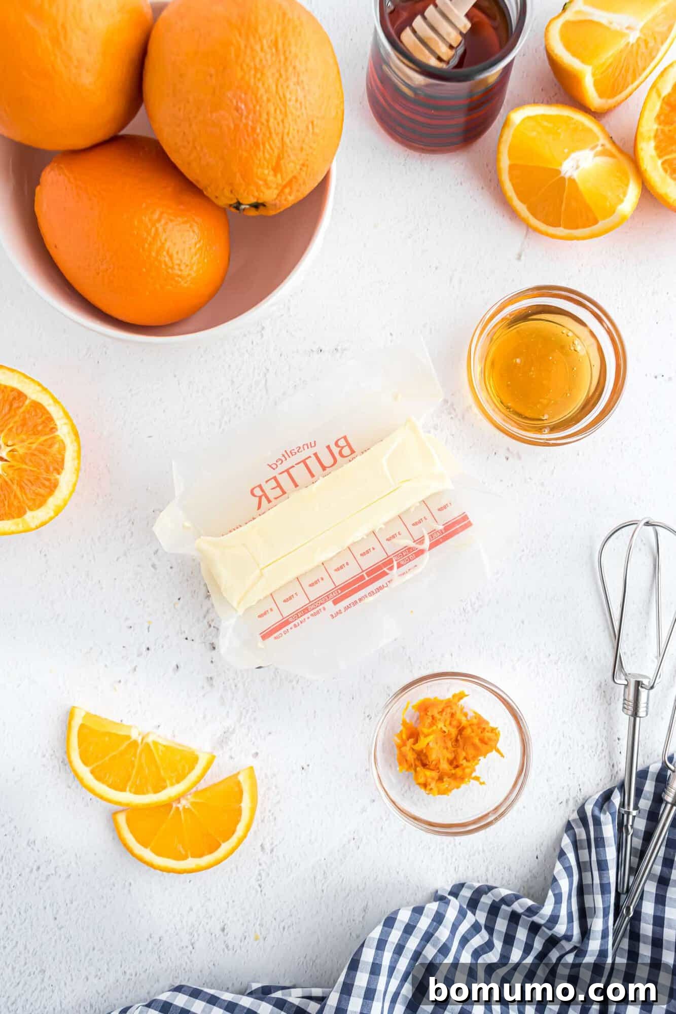 An overhead view of the fresh ingredients laid out for making orange honey butter: a stick of softened butter, a whole orange, and a jar of golden honey.