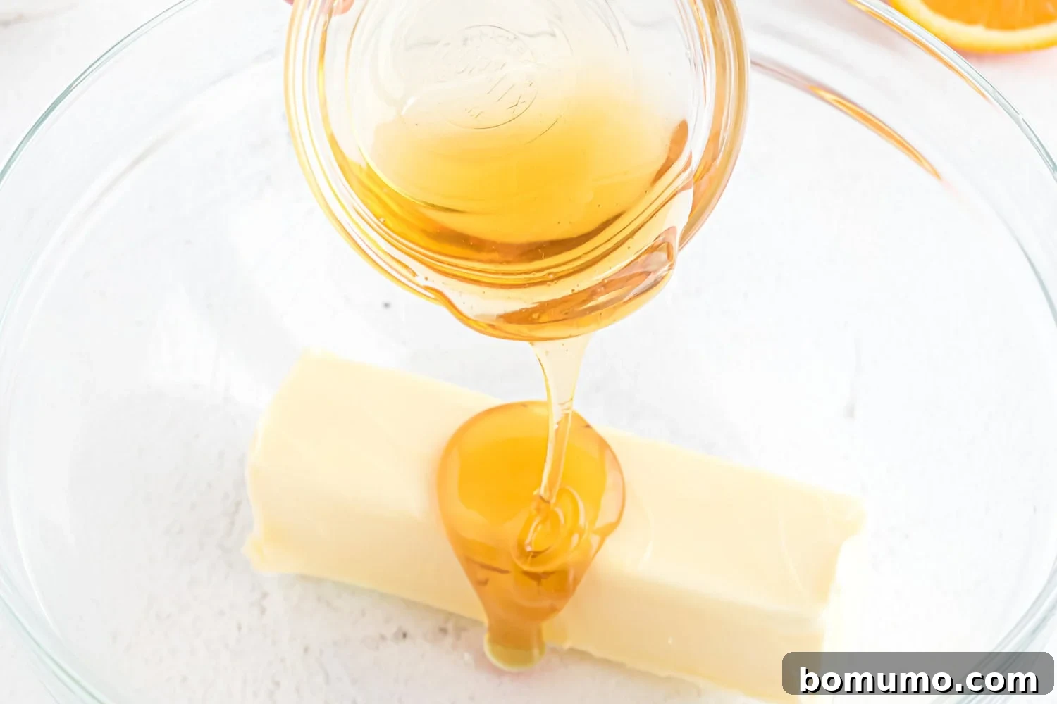 Smooth golden honey being drizzled over softened butter in a clear glass bowl, ready for blending.