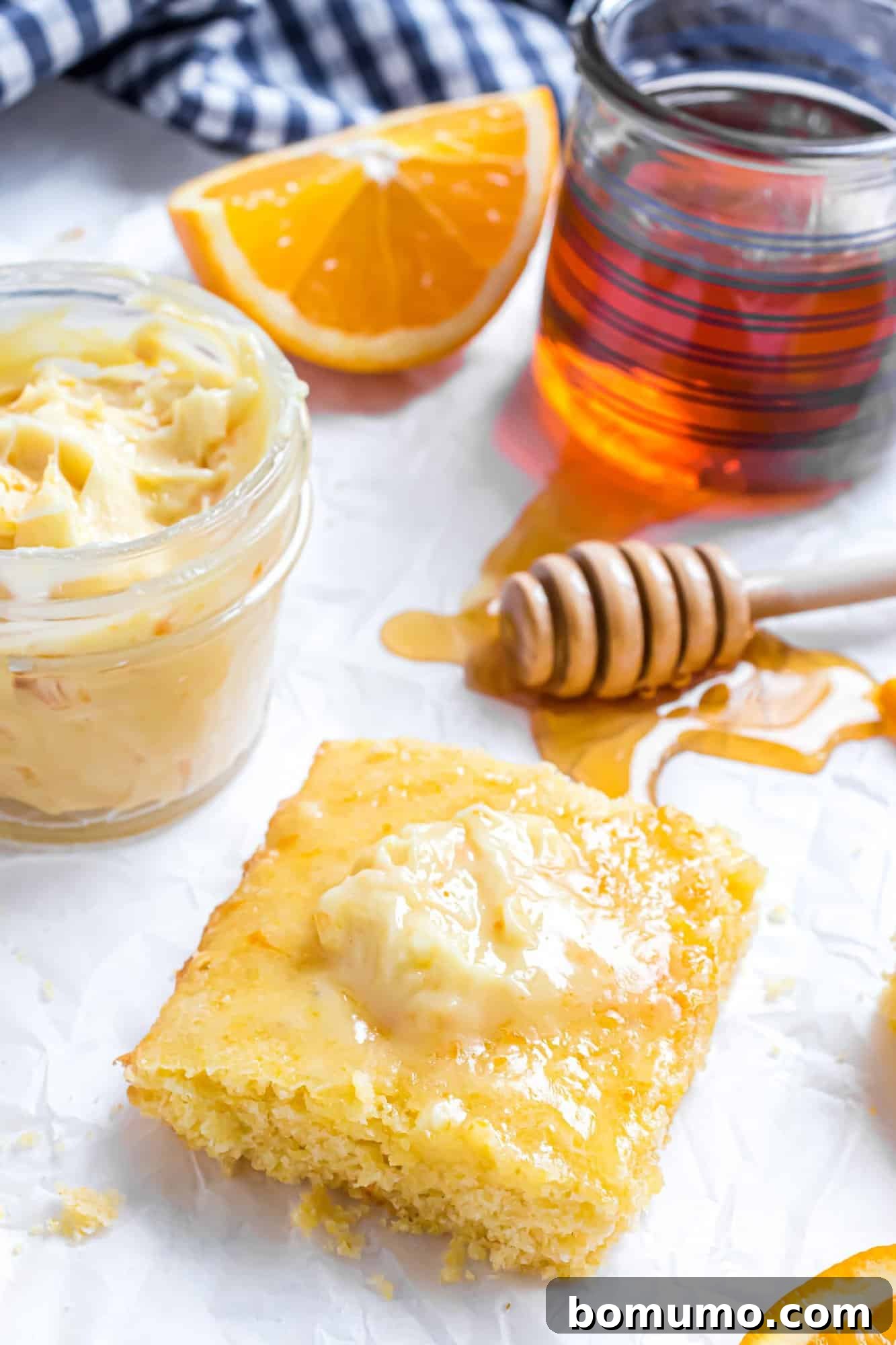 A slice of warm cornbread topped generously with creamy orange honey butter, with a small jar of honey blurred in the background, creating a rustic and inviting scene.