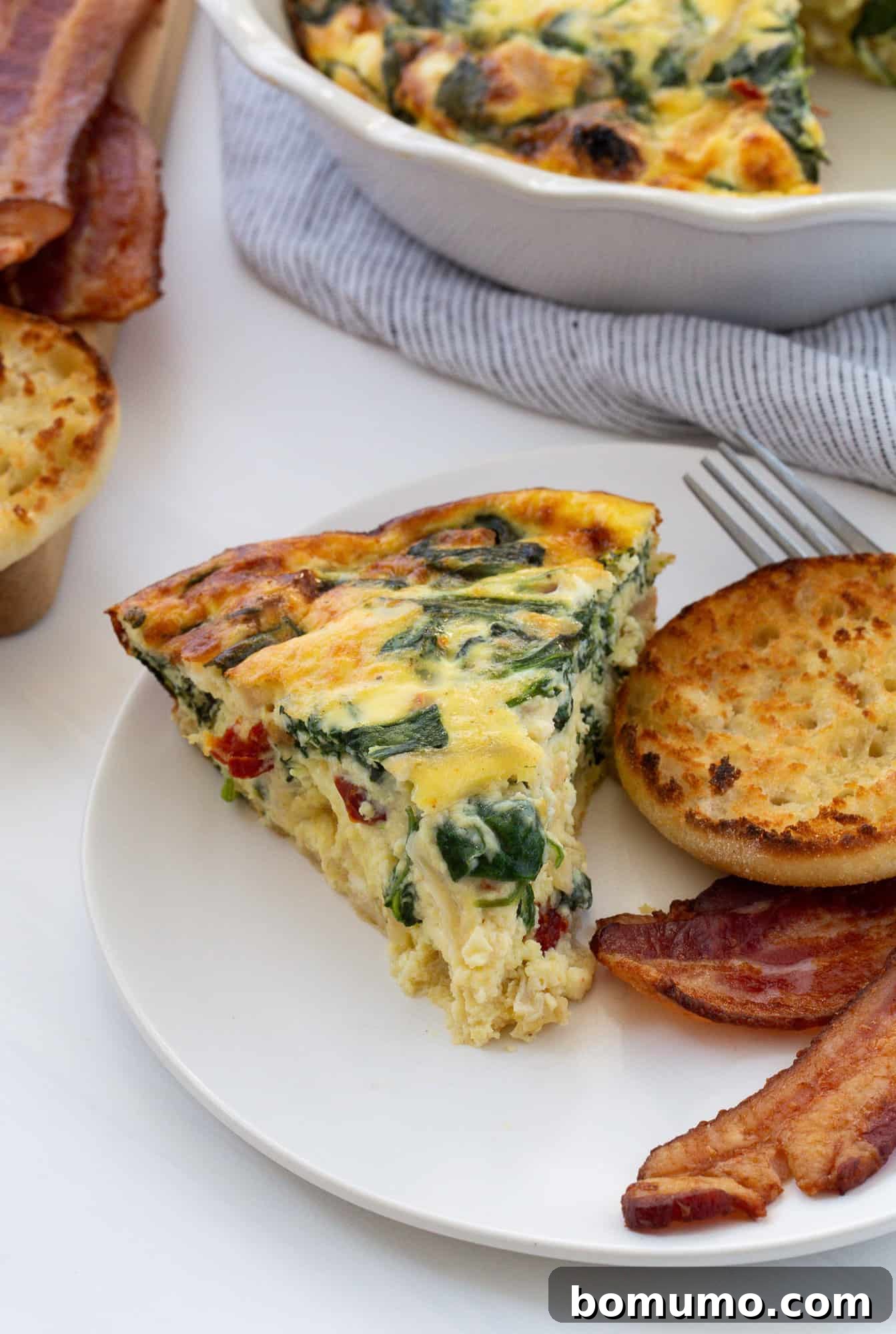 A delectable slice of crustless quiche, generously filled with spinach, caramelized onions, sun-dried tomatoes, and cheese, resting on a white plate. It's accompanied by a golden English muffin and crispy bacon, set against a rustic background, showcasing a perfect brunch spread.