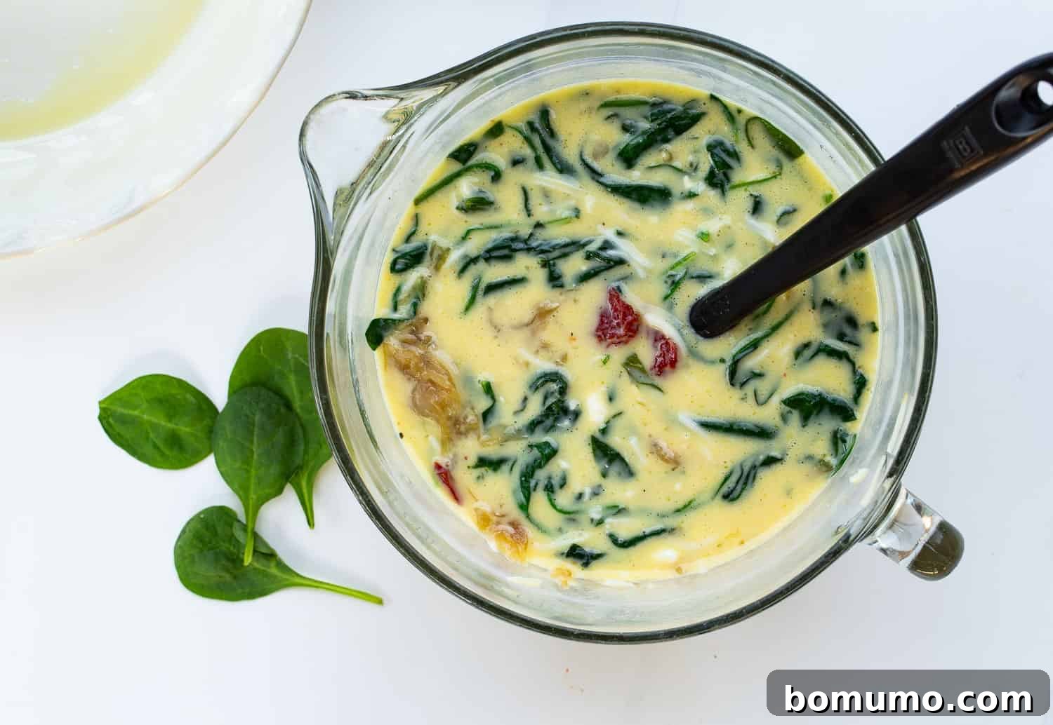 All the chosen add-ins – cooked spinach, caramelized onions, sun-dried tomatoes, mozzarella, and feta cheese – stirred into the egg mixture in a large bowl, creating a colorful and rich filling.
