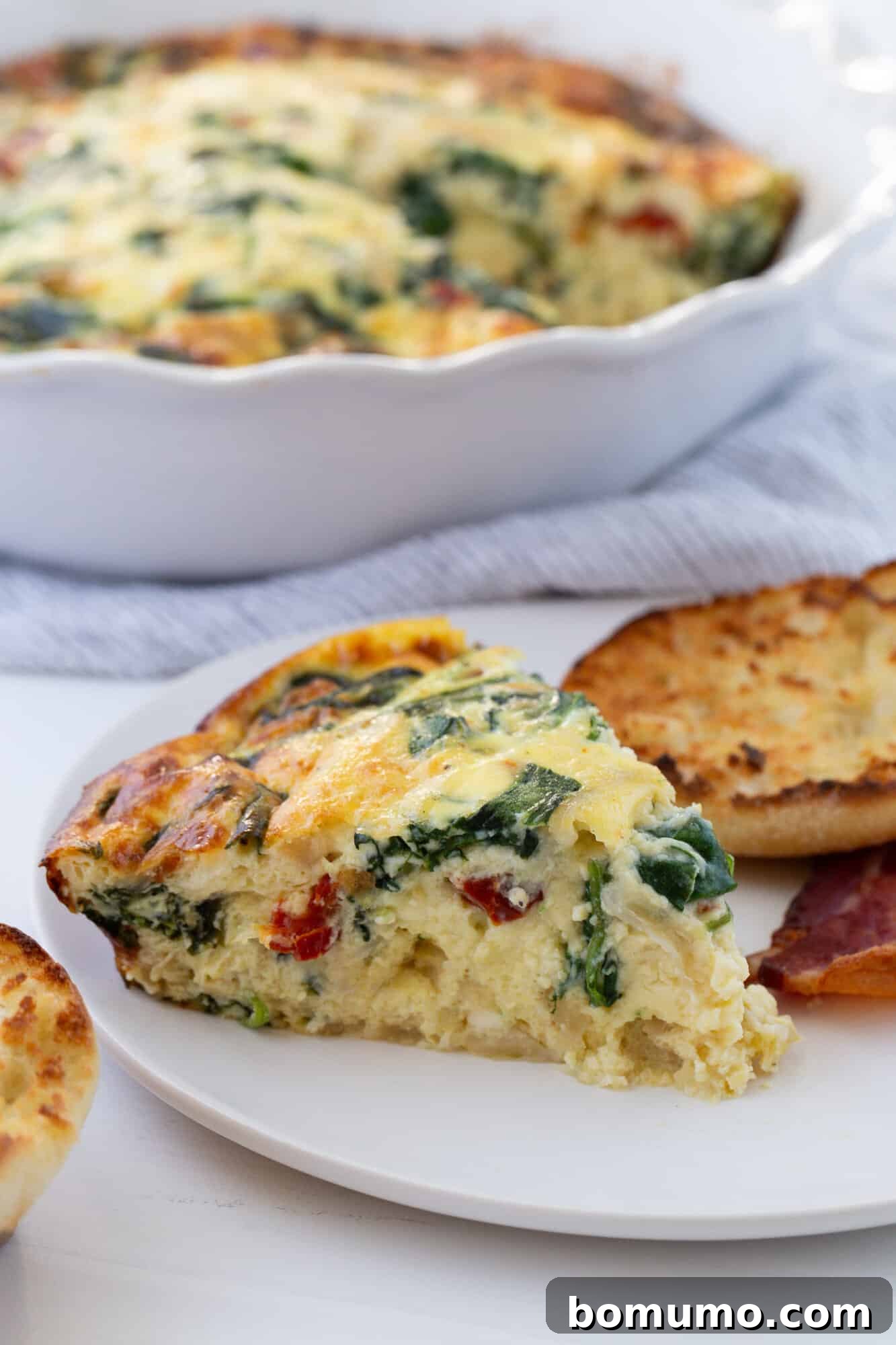 A perfectly portioned slice of crustless quiche, showcasing its layers of filling, presented on a white plate next to a toasted English muffin, ready for serving.