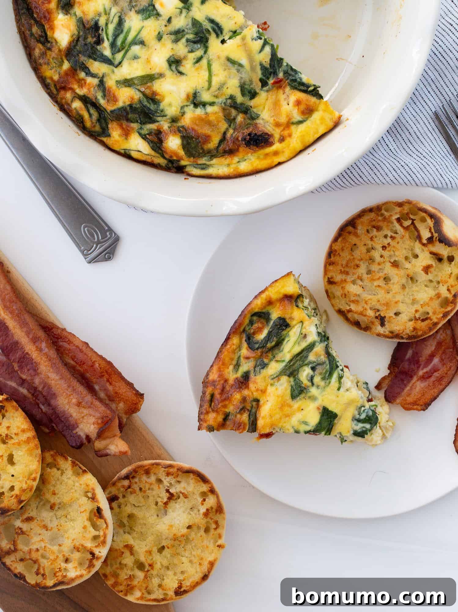 An overhead shot of a freshly baked crustless quiche with a slice removed, revealing its vibrant green spinach, golden onions, and red sun-dried tomatoes within the fluffy egg custard. It's served alongside a toasted English muffin, ready for a delightful breakfast or brunch.