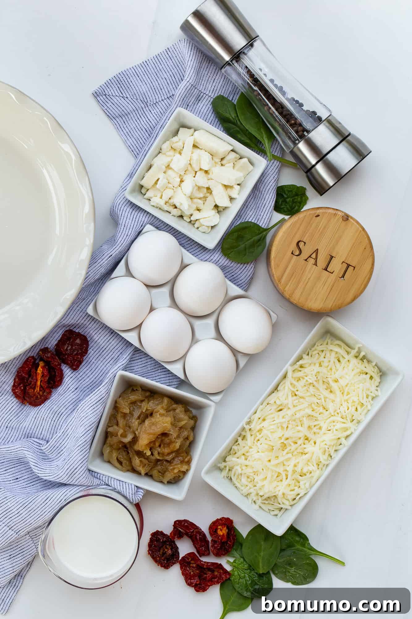 A flat lay photograph showcasing all the fresh and vibrant ingredients neatly arranged: eggs, whole milk, spinach, sun-dried tomatoes, caramelized onions, mozzarella, feta, and various seasonings, ready for making the crustless quiche.