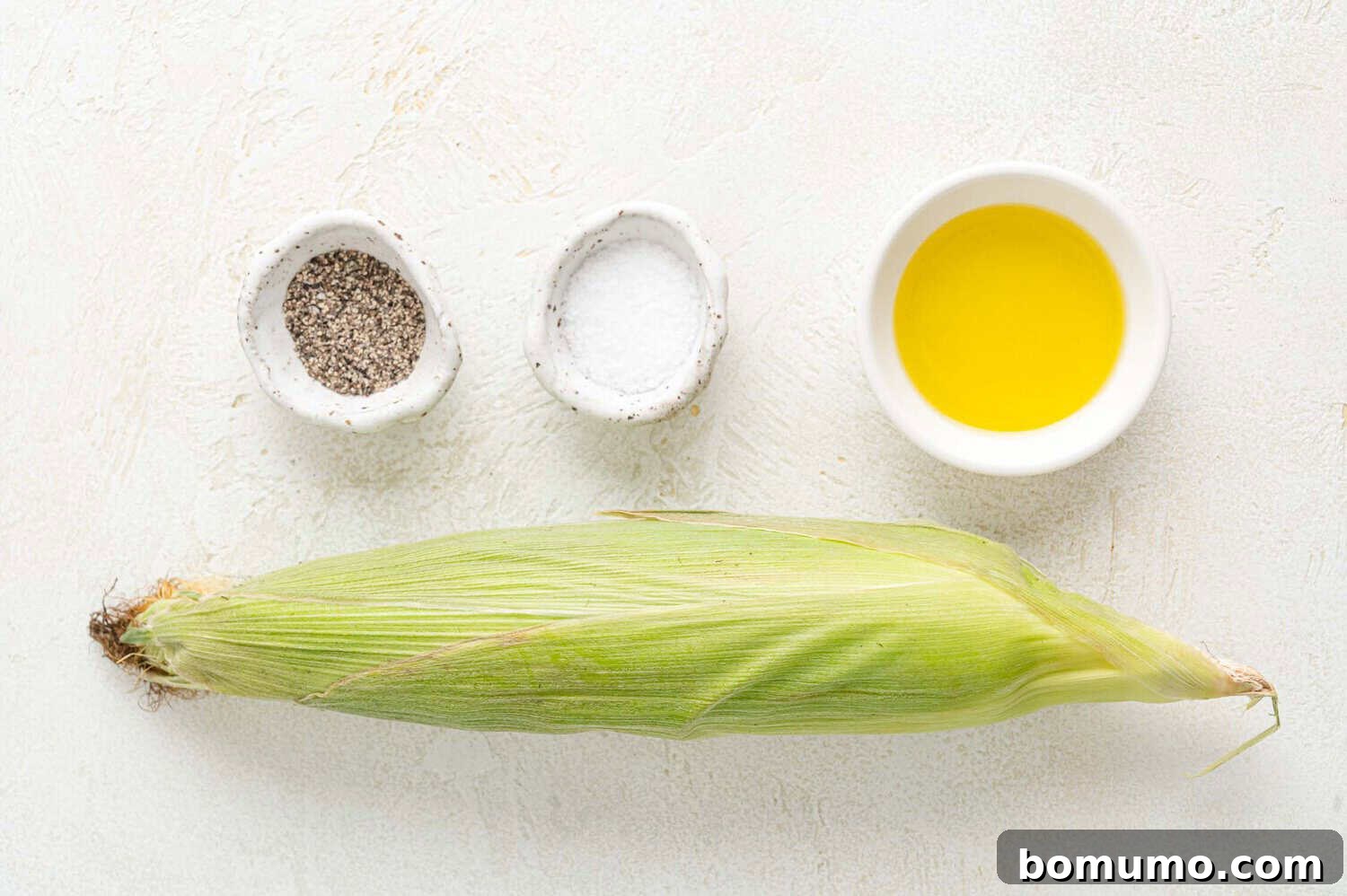 An ear of corn in husk is placed on a white surface with small bowls of black pepper, salt, and olive oil arranged above it, demonstrating ingredients.